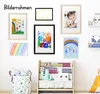 A collection of framed children's artwork and a photo of a child with a dog displayed on a white wall above a wooden shelf unit filled with toys and decorative items. Produktbild-Vorschau