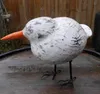A wooden bird sculpture with an orange beak and black wire legs stands on a wooden surface in front of a wooden fence. Produktbild-Vorschau