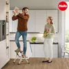 A couple in a modern kitchen, the man is standing on a step stool reaching for something on a high shelf, the woman is holding flowers. Náhled obrázku produktu