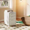 A white filing cabinet with three drawers sits next to a window, with books and a cupcake on top. The floor has a patterned rug, and a large brown beanbag chair with a green blanket is visible on the right. Náhled obrázku produktu