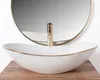 A white oval basin with a gold rim sits on a wooden surface, with a gold faucet and a round mirror in the background. Anteprima dell’immagine prodotto