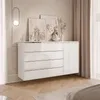 A modern white dresser with four drawers and a cabinet is placed against a textured wall, adorned with artwork and a decorative lamp. The scene is completed with draped curtains and a wooden floor. Produktbild-Vorschau