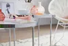 A close-up of two round nesting tables with glass tops and chrome legs, decorated with vases, a notebook, and a ribbon. Produktbild-Vorschau
