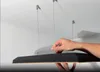 A hand is shown adjusting the suspension of a black acoustic ceiling panel. The panel is suspended from the ceiling by thin wires. Podgląd zdjęcia produktu