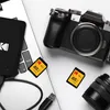 A collection of photography equipment including a camera, lens, SD cards, and a portable hard drive on a laptop. Produktbild-Vorschau