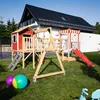 A wooden playground set with a red playhouse, swings, and a slide is set up on a green lawn in front of a modern house. Produktbild-Vorschau