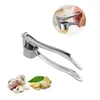 A stainless steel garlic press with a white handle, shown on a white background. There are also some garlic bulbs and ginger slices in the lower left corner. A small inset image shows the garlic press in use. Náhled obrázku produktu