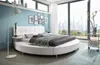 A modern bedroom with a large round bed, floor lamp, and a city view through the window. Produktbild-Vorschau