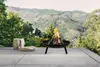 A fire pit is shown on a patio with a scenic mountain and forest background. Produktbild-Vorschau