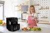 A woman in a kitchen holding a tablet, with an air fryer and food on the counter. Podgląd zdjęcia produktu
