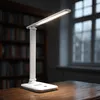 A white LED desk lamp with a Qi charger is placed on a wooden desk in front of a bookshelf and a window. Produktbild-Vorschau