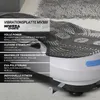 A close-up, angled view of a white and blue vibration plate with black textured surface, set against a blurred background of a rug and floor. Produktbild-Vorschau
