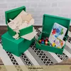Two green wooden boxes with lids, one open and filled with wooden craft shapes and the other open and filled with paints and a notebook, placed on a patterned rug. Produktbild-Vorschau