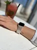 A person's wrist wearing a silver watch is shown in the foreground, with a blurred drink in the background. Produktbild-Vorschau