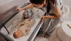 A woman is adjusting a baby in a crib next to her bed. Produktbild-Vorschau
