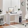 A white computer desk with a monitor, keyboard, lamp, and storage shelves is set up in a room with blinds and a clock on the wall. Produktbild-Vorschau