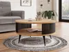 A round coffee table with a wooden top and black metal slatted sides is placed on a circular rug in a living room setting. A grey sofa is visible to the left, and a large window with greenery outside is to the right. Náhled obrázku produktu