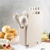 A kitchen utensil set with a cutting board is displayed on a countertop with bread rolls and a bottle of oil in the background. Produktbild-Vorschau