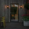 An outdoor wall lamp is mounted on a wooden wall next to a glass door, with outdoor furniture and plants visible. Produktbild-Vorschau