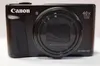 A black Canon PowerShot digital camera is shown from the front against a white background. Podgląd zdjęcia produktu