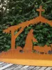 A metal nativity scene with a cross on top, set against a backdrop of green foliage. Anteprima dell’immagine prodotto