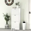 A white shoe cabinet with three flip-down drawers is shown in a hallway next to a door. A large clock hangs on the wall above the cabinet, and a potted plant stands to the left. A white umbrella stand with umbrellas is to the right of the cabinet. Produktbild-Vorschau