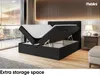A bedroom scene with a bed that has its storage compartment open, revealing the mattress and topper. The bed is upholstered in black fabric and is set against a wall with vertical wooden slats. A bedside table with a lamp is visible to the right. Produktbild-Vorschau