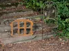 An old metal window frame and a metal chair are placed on stone steps outdoors, surrounded by greenery and fallen leaves. Produktbild-Vorschau