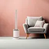 A white tower fan is placed next to a gray armchair in a room with a pink wall. Podgląd zdjęcia produktu