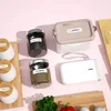 A collection of spice jars, a lunchbox, and a label maker are arranged on a pink surface, with some fresh herbs and a red pepper in the foreground. Podgląd zdjęcia produktu