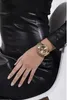 A close-up shot of a woman's wrist wearing a gold Guess watch, with her hand resting on a black leather garment. Náhled obrázku produktu