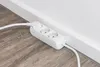 A white power strip with three outlets and a white cable is placed on a wooden floor next to a white baseboard and wall. Produktbild-Vorschau