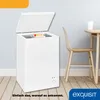 A white chest freezer is shown with its lid open, displaying some frozen food items. It is placed on a wooden floor next to a table and chair in a room with light gray walls. Produktbild-Vorschau