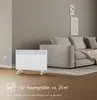 A white electric heater is positioned on a wooden floor in front of a gray sofa. There is text overlay on the bottom left corner. Podgląd zdjęcia produktu