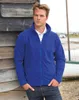 A man wearing a blue fleece jacket and jeans, leaning against a wooden wall outdoors, with a boat in the background. Produktbild-Vorschau