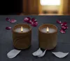 Two LED candles in wooden holders are placed on a dark surface, surrounded by scattered pink and white flower petals. The background is softly blurred, suggesting an indoor setting with warm lighting. Voir les images du produit