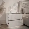 A white, high-gloss nightstand with two drawers, placed next to a bed. A table lamp and a bust are on top of the nightstand. Produktbild-Vorschau