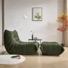 A green armchair and ottoman are placed in a living room setting with a floor lamp, a small side table with a vase and books, and a decorative plant. Produktbild-Vorschau