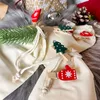Christmas-themed wooden clothespins with tree and mitten designs are attached to small drawstring bags, with a pine cone and gift wrapping in the background. Produktbild-Vorschau