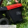 A person is holding the lid of a lawnmower's grass catcher bag, which is filled with grass clippings. The lawnmower is in a natural outdoor setting with greenery and foliage. Produktbild-Vorschau