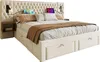 A beige upholstered bed with a tufted headboard, integrated bedside tables with lights, and storage drawers underneath. Two tablets are placed on the headboard. Náhled obrázku produktu