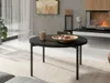 A modern living room scene with a round black coffee table in the foreground, featuring a mug and a bowl of cookies. A fireplace with a fire is visible in the background. Produktbild-Vorschau
