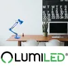 A blue desk lamp is placed on a wooden desk in front of a white wall. A calendar and a potted plant are visible in the background. The LUMILED logo is at the bottom. Náhľad obrázku produktu