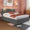 A bedroom scene featuring a bed with a grey upholstered headboard and base, adorned with an orange throw blanket and pillow. The room has pink walls and wooden flooring. Produktbild-Vorschau
