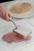 A hand is shown using a meat tenderizer on a piece of raw meat, which is covered in plastic wrap. In the background, a breaded cutlet is on a plate with breadcrumbs. Podgląd zdjęcia produktu