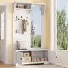 A white entryway furniture piece with a bench, storage cabinets, hooks, a mirror, and a shelf, placed in a room with a window showing greenery. Produktbild-Vorschau