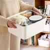 A person is holding a white storage box filled with various food items and a bowl. The box is placed on a shelf with books and other storage containers. Produktbild-Vorschau
