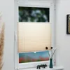 A cream-colored pleated blind is installed on a window, with a blurred view of greenery and flowers outside. Decorative items are placed on the windowsill. Produktbild-Vorschau