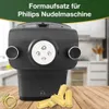 A black pasta maker attachment sits on a wooden table with some pasta tubes in front of it. The background is a blurred kitchen scene. Produktbild-Vorschau