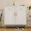 A white sideboard with gold legs and handles, featuring two drawers and two doors, is shown in a room setting. An umbrella stand with an umbrella is to the left, and slippers are on the floor in front. A diffuser and a small radio are on top.